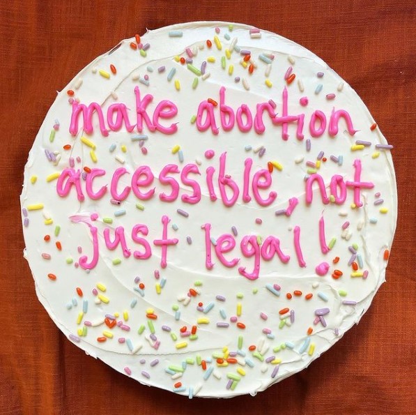 Round cake with white frosting and rainbow sprinkles, with pink icing text that says make abortion accessible, not just legal, from the DC Abortion Fund.