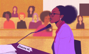 A person with medium brown skin and glasses, wearing a suit, sits in front of a microphone, giving testimony at a legislative hearing. In the gallery seats behind them, a number of people listen attentively.