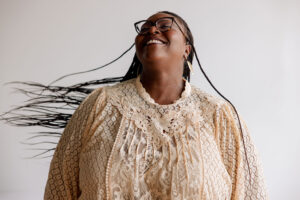 NNAF Executive Director Oriaku Njoku wears a beige shirt and stands in front of a white background. They are smiling and shaking their long braids.