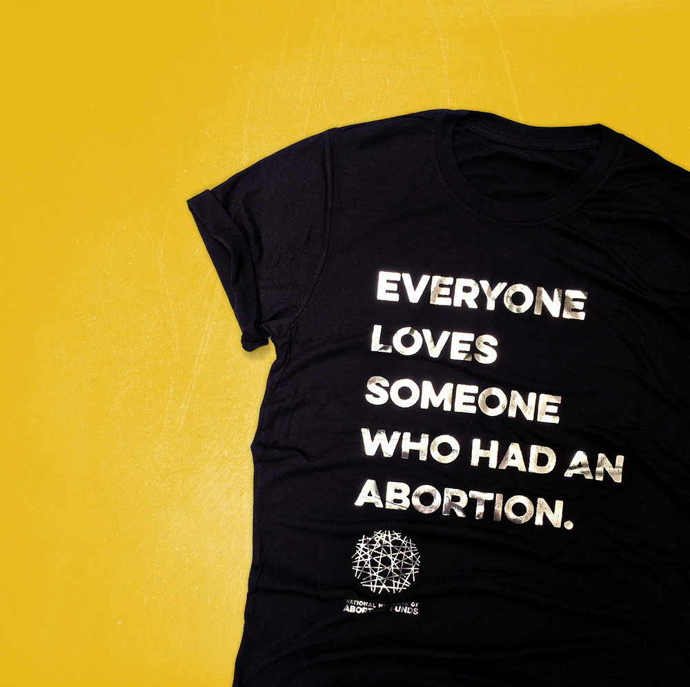 Una camiseta negra colocada en ángulo sobre un fondo amarillo mostaza. Letras plateadas metálicas en el frente de la camiseta dicen EVERYONE LOVES SOMEONE WHO HAD AN ABORTION (en inglés) en mayúsculas sobre el logo de NNAF.